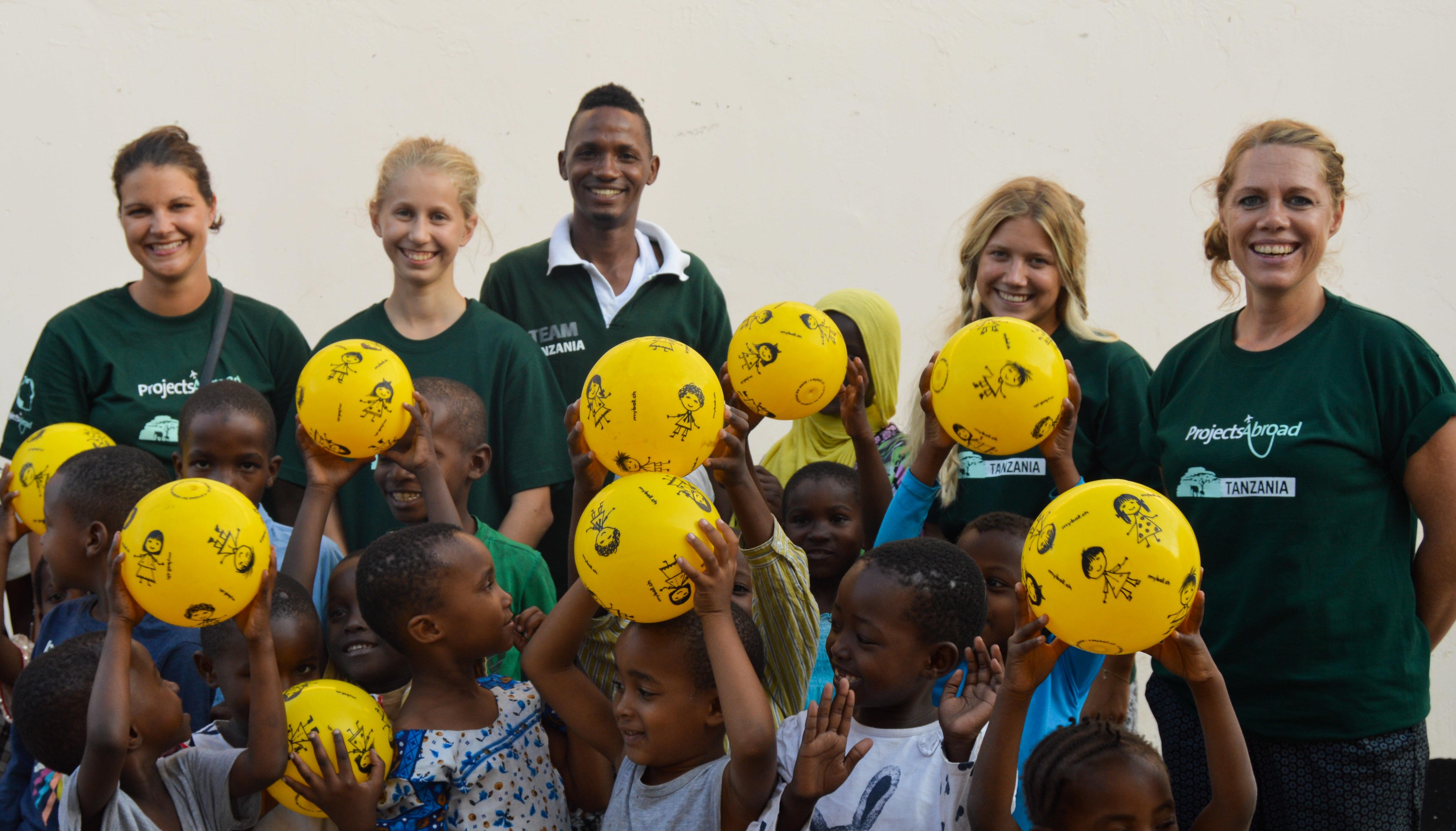 Projects Abroad volunteers and staff pose for a picture with children holding yellow balls above their head during a Care Outreach in Tanzania. Childcare volunteers working with children in Tanzania pose for a picture with teachers and children during a Care outreach.